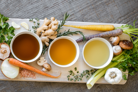 three cups of broth on a wooden board surrounded by garlic, carrots, ginger and mushrooms