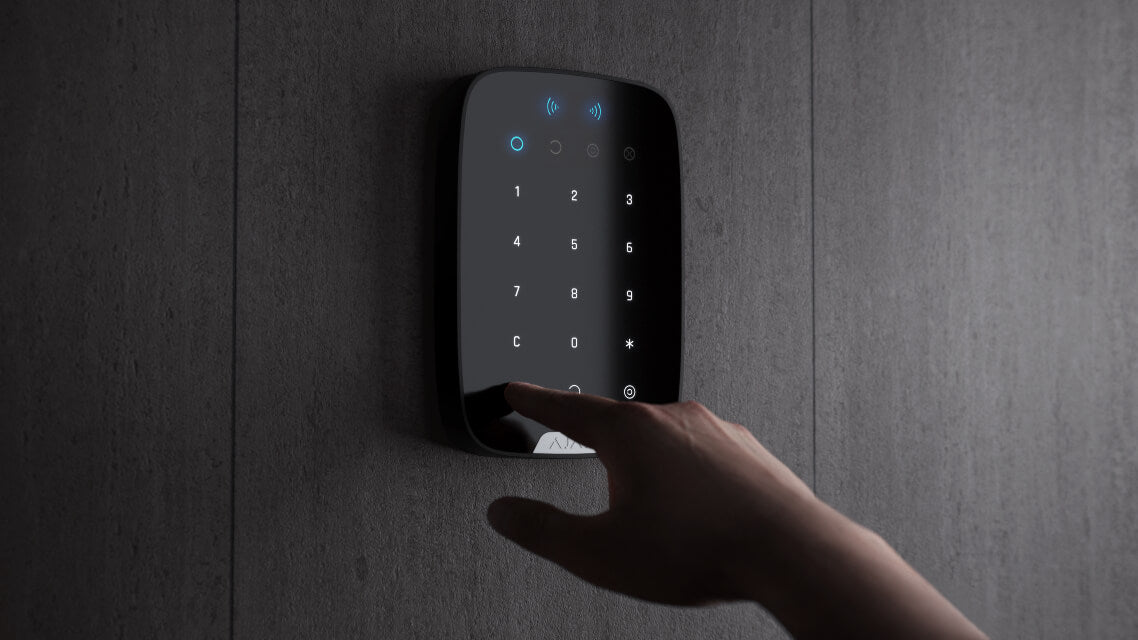 A Black Ajax Security Systems Keypad Plus mounted on a wall, with a hand reaching out to touch the touch keypad.