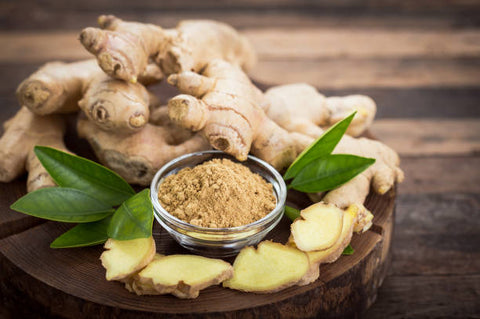 Small pile of ginger root on a table with a glass container of ground ginger in front of it.