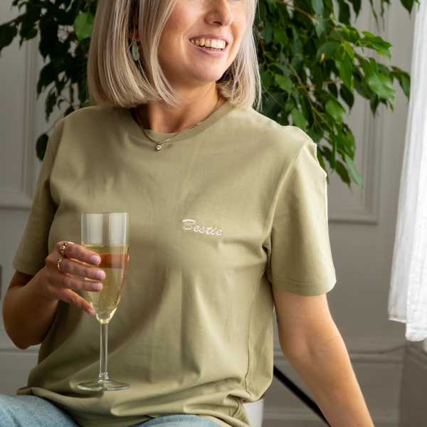 woman wearing a sage green cotton t-shirt embroidered on the breast pocket with the word "bestie" in cream