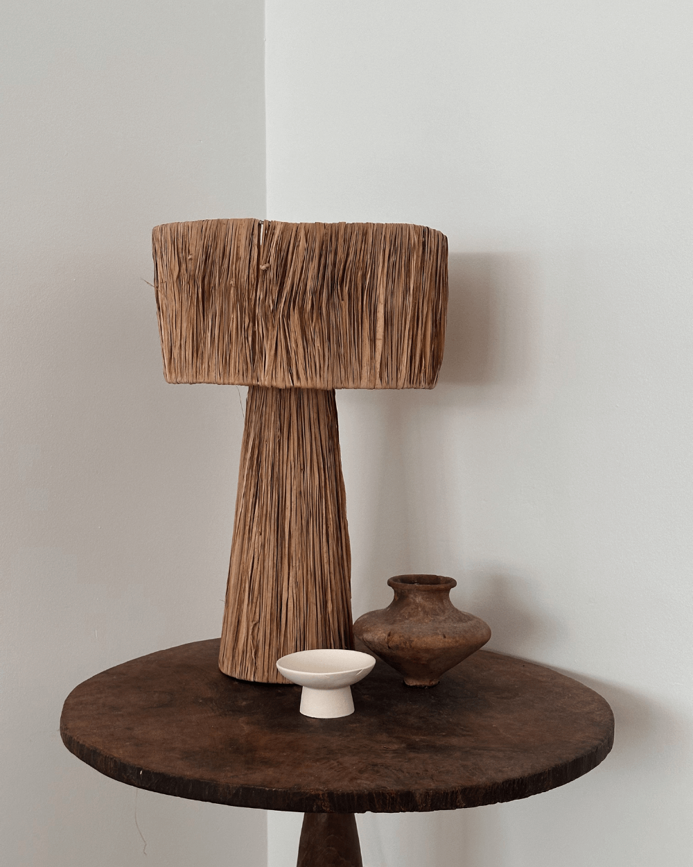 Natural fiber lamp with a woven shade on a rustic wooden table, accompanied by a small white dish and a dark pottery vase, set against a light-colored wall.
