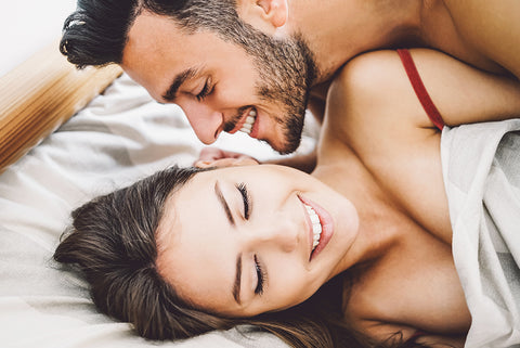 couple laying in bed together with the man on top of the women looking at her lovingly and playfully the women with brown hair is smiling to herself