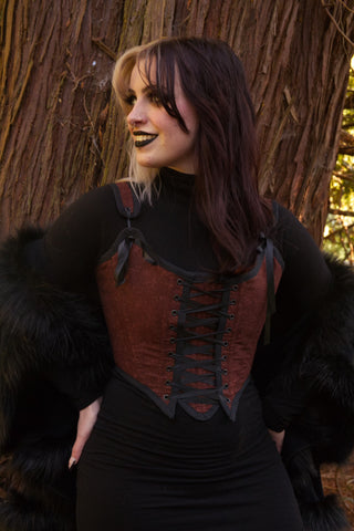 femme presenting person in front of tree wearing black dress and dark red bodice.