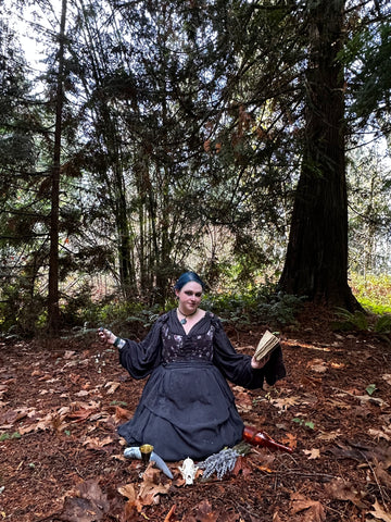 woman in black dress and floral bodice sitting on fall leaves in forest holding charms and book