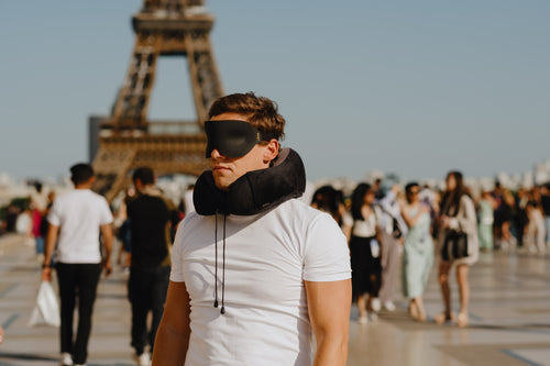Grunnleggeren står smilende utenfor Eiffeltårnet i Paris, iført en sovemaske og en nakkepute, og representerer en kombinasjon av reisekomfort og kjente landemerker.