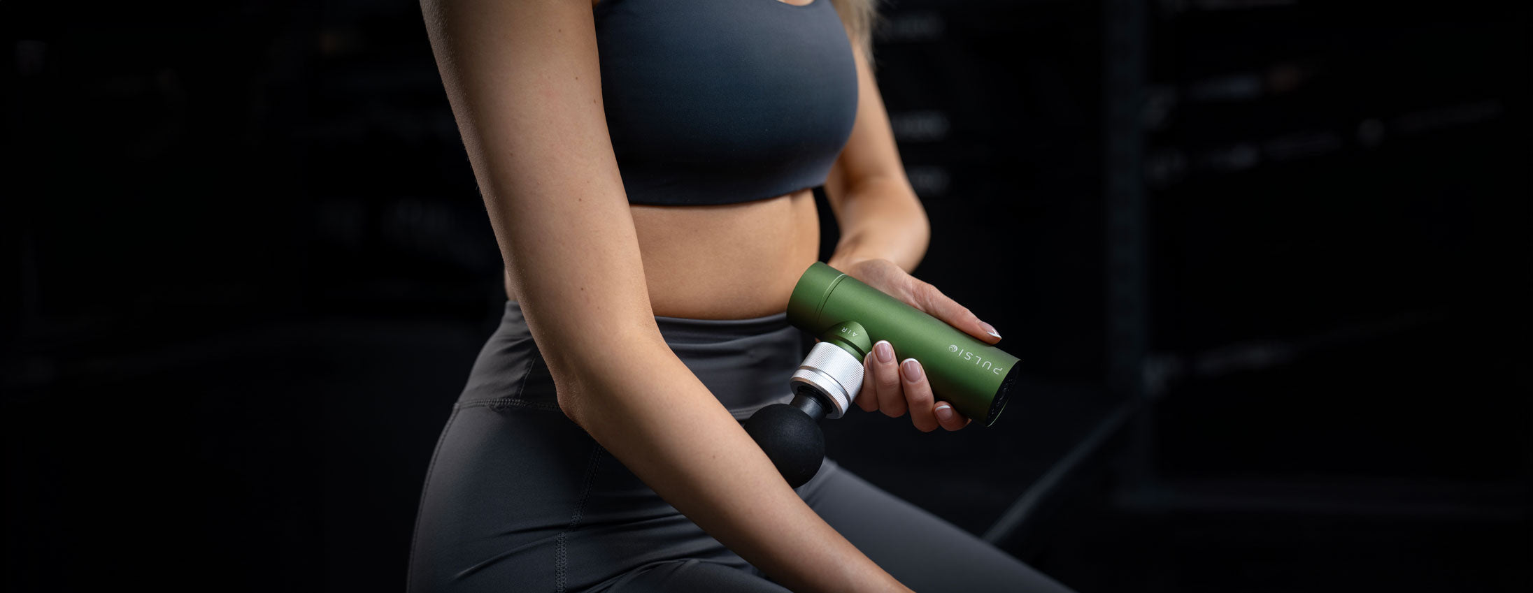 Woman holding a massage gun, focused on abdominal muscles, dark gym background.