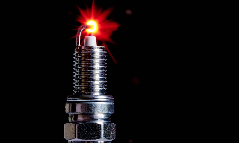 A close-up profile of a nickel-plated spark plug with a spark at the end of the electrode against a black background.