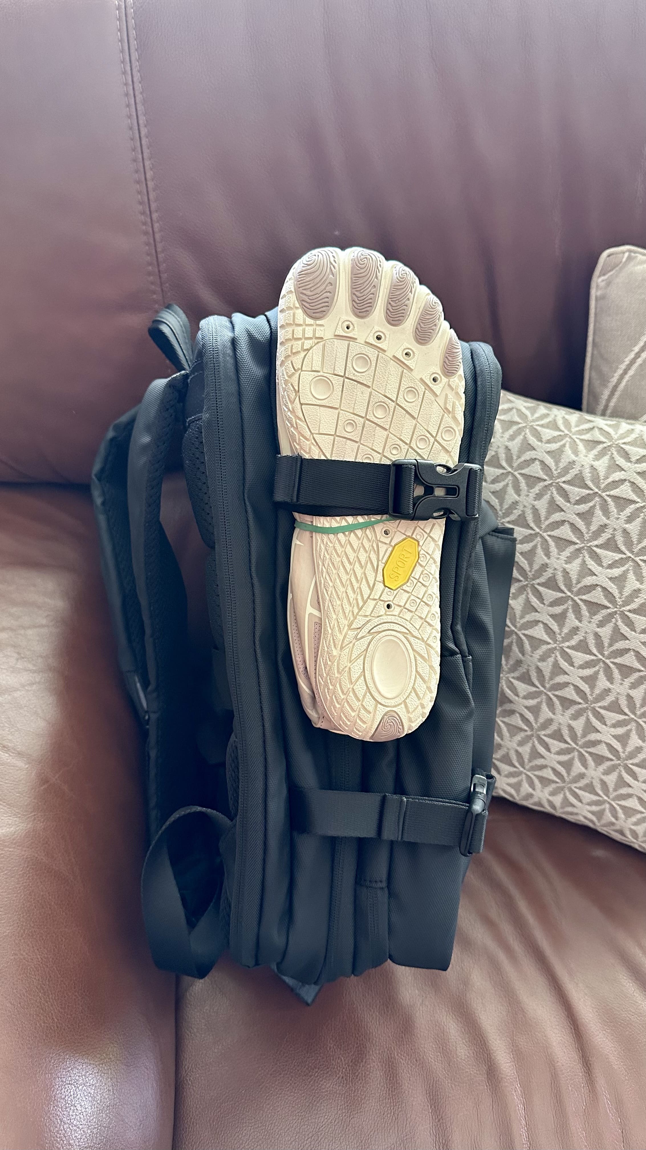 A black backpack with a strap holding a white shoe, placed on a leather couch.