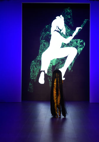 Runway model showcasing Johny Dar's body art, with floor-length hair and a backdrop of Dar's artwork displayed on an LED screen.