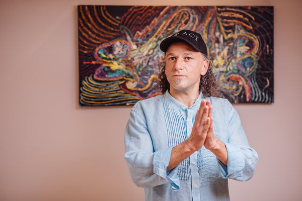 Artist Johny Dar standing before one of his paintings, hands folded in front, wearing an AOI hat.