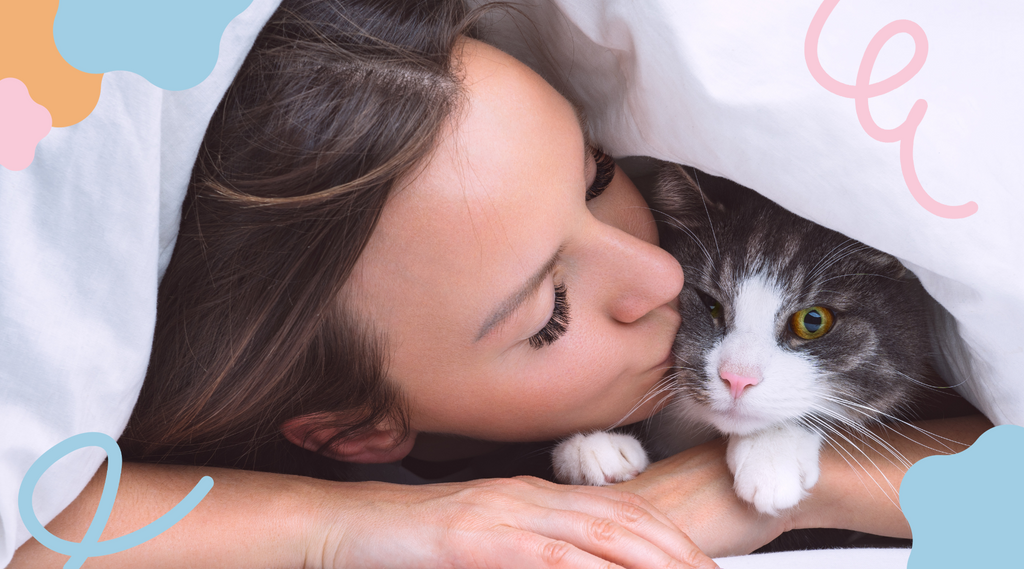 An image of a woman affectionately kissing a cat nestled under sheets, sharing a tender moment of closeness and warmth.