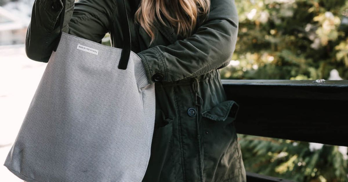 A person in a green jacket holding a stylish houndstooth tote bag, standing by a railing outdoors, focus on the bag.