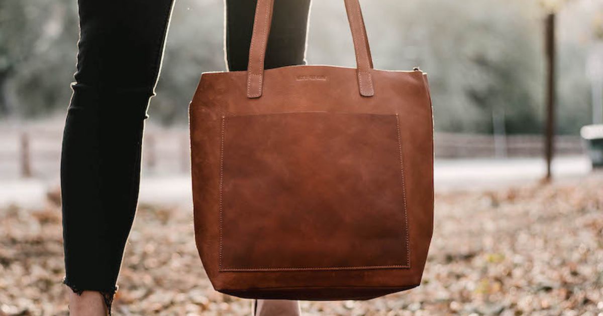 Close-up of a person holding a large brown leather tote bag, standing in a park with fallen leaves, focused on the bag with the background softly blurred.