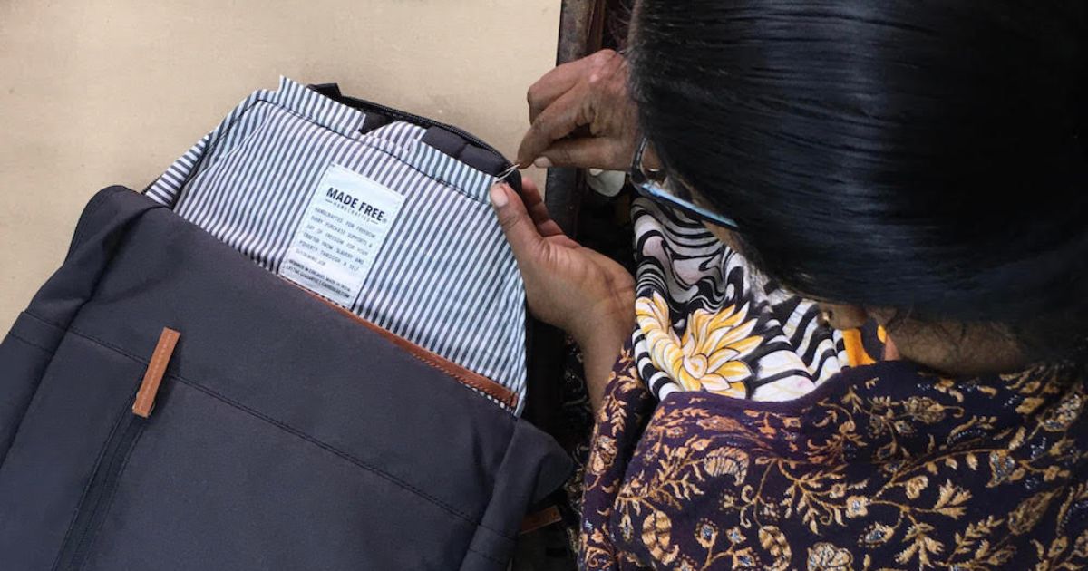 A woman with dark hair is seen from above, sewing a label into a striped lining of a gray backpack. The label reads "HANDMADE".