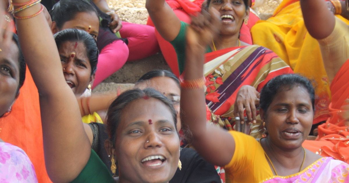 A group of joyful women dressed in colorful traditional Indian attire, celebrating with raised hands and smiles at an outdoor event.