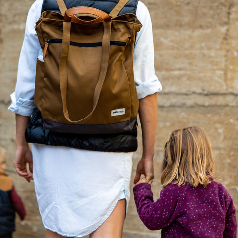 A person wearing a white dress and a brown backpack walks hand-in-hand with a child in a purple jacket. The child has blonde hair and looks towards another child in the background. The group is outdoors in front of a textured stone wall.