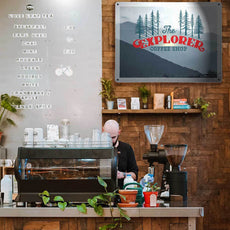 A barista in a cafe with a large magnetic board on a wooden wall behind the counter with a logo sign that reads 'The explorer' coffee shop