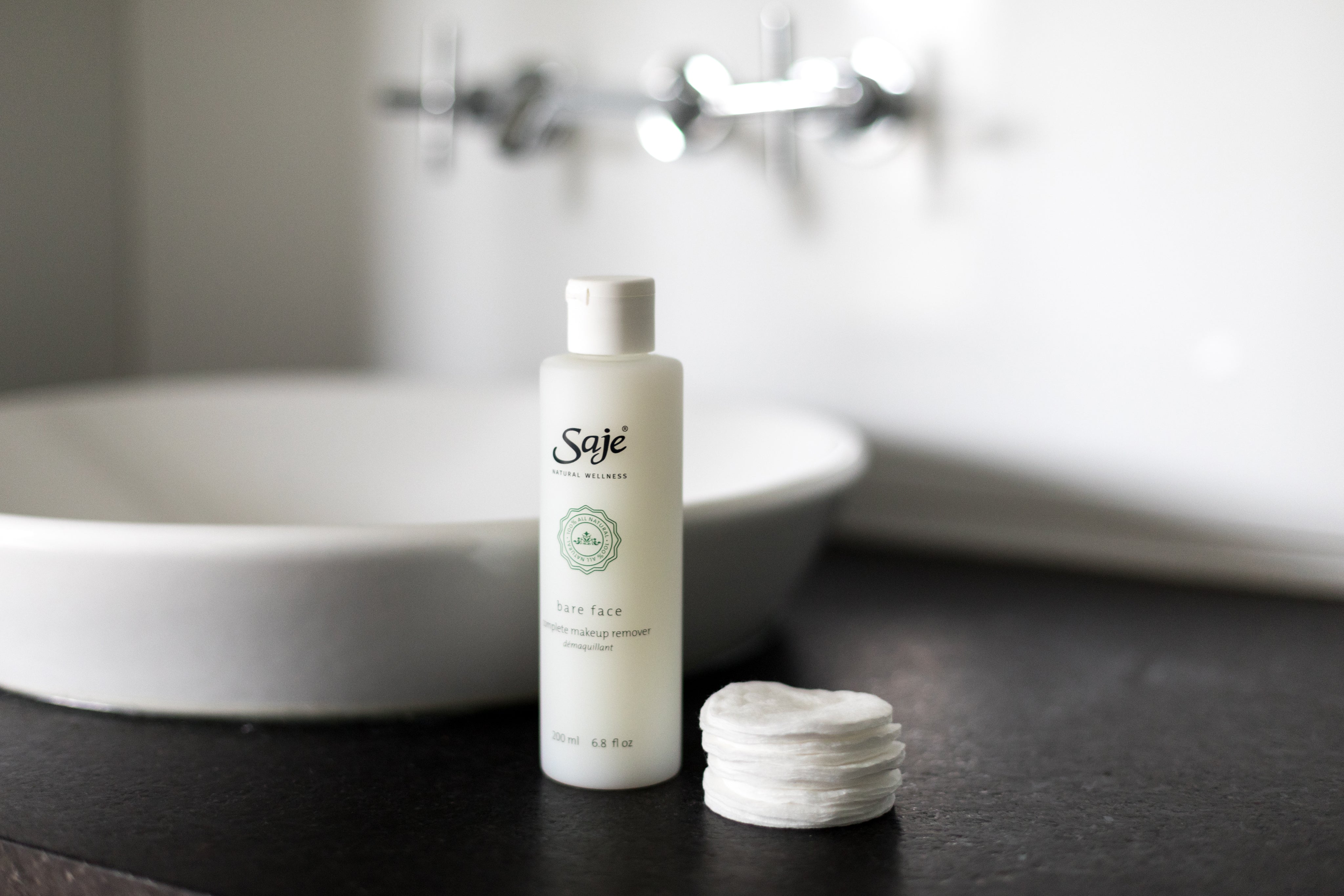 Saje bare face makeup remover on a bathroom counter next to a stack of round cotton pads