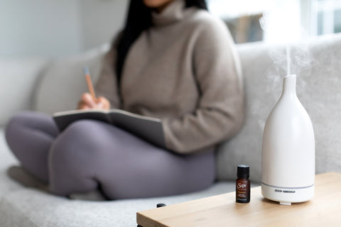A person journaling on a couch with a Saje diffuser sitting on a table beside them