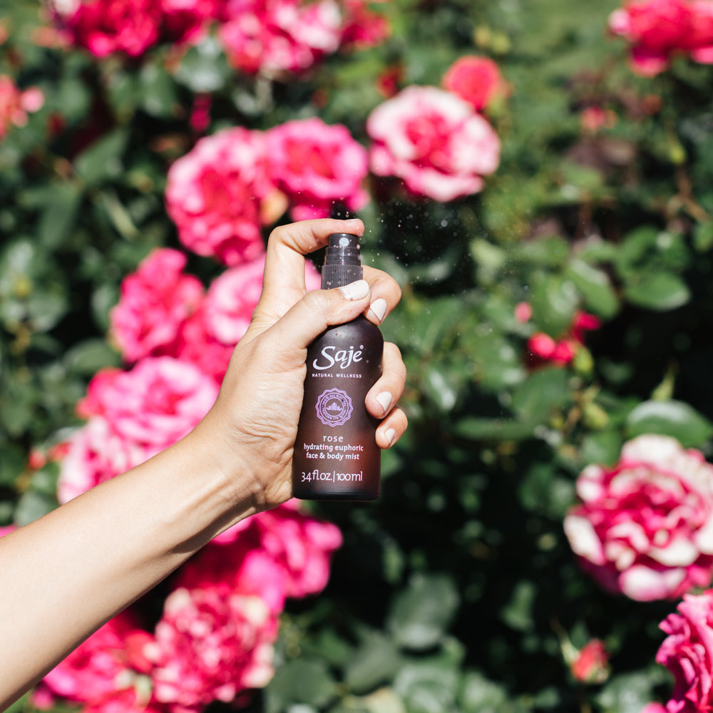 A person'd hand spraying rose mist in front of pink roses