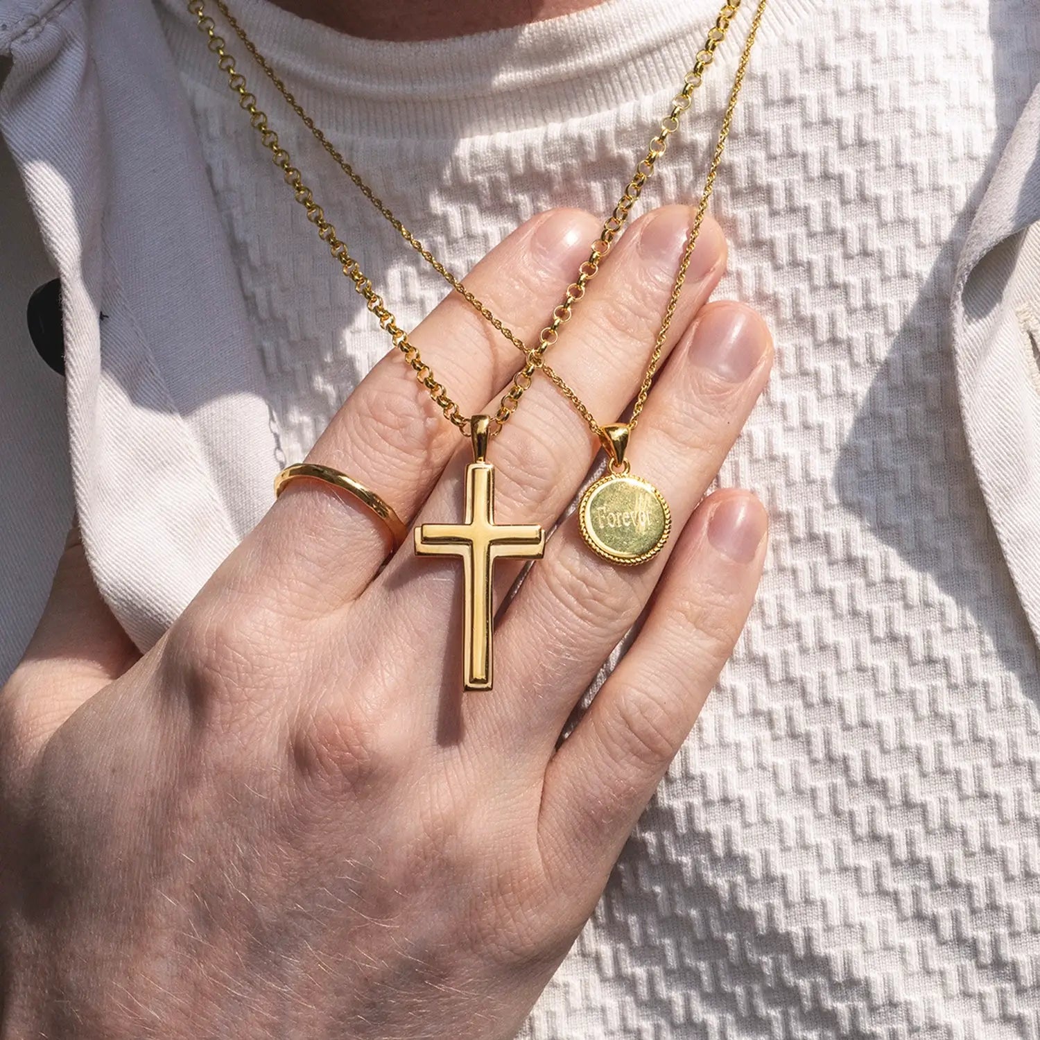 Close up of male model in cream textured crew neck top showing off a heavy gold cross necklace and a gold engravable disc necklace by Lily Blanche by draping them on the back of his hand