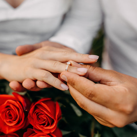 Die Hand des Bräutigams zieht der Hand der Braut einen Verlobungsring behutsam über den Finger. Unter den beiden Händen ist ein Strauß mit roten Rosen.