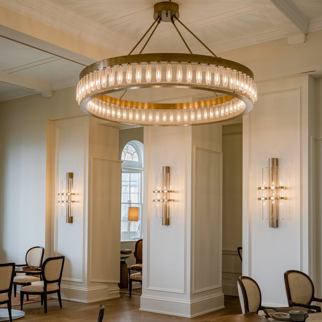 A photo of a room with a bespoke lighting design. There is a large circular chandelier made of brass and glass in the centre of the room. There are also wall sconces with brass and glass fixtures on either side of the room. The room has a high ceiling and the walls are painted white.