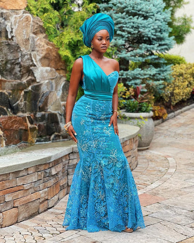 A young woman stands outdoors, exuding elegance in a teal blue Aso Ebi gown made by Senami Atinkpahoun of Je Suis NYC with intricate lace detailing. The gown flares gently at the bottom, creating a mermaid silhouette. She is adorned with a beautifully wrapped Gele head tie in the same vibrant hue, complementing her dress. Her expression is contemplative and poised against a backdrop featuring a water feature and lush greenery.