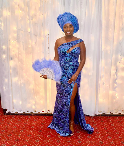 A woman stands confidently in front of a white curtain with twinkling lights, wearing a striking blue Aso Ebi dress made by Senami Atinkpahoun of Je Suis NYCwith metallic patterns and a high slit. She complements her attire with a matching blue head wrap and holds a fluffy blue fan. Her poised demeanor and radiant smile reflect the festive spirit of the occasion.