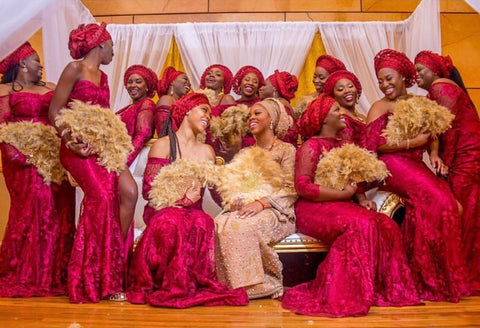 A joyful group of women at a celebration, all dressed in coordinating deep red Aso Ebi attire with intricate lace detailing and matching head wraps. They are holding fluffy, light brown decorative fans and are gathered around a seated woman in a golden, embellished dress, sharing a moment of laughter and camaraderie.