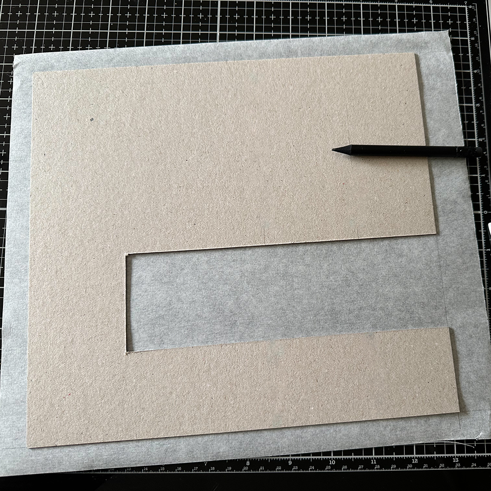 Eine graue Pappe mit einem E-förmigen Ausschnitt liegt auf einem transparenten Bogen Papier, der wiederum auf einer schwarzen Schneidematte mit weißen Gitternetzlinien platziert ist. Ein schwarzer Bleistift zeigt auf den oberen Rand des Ausschnitts in der Pappe.