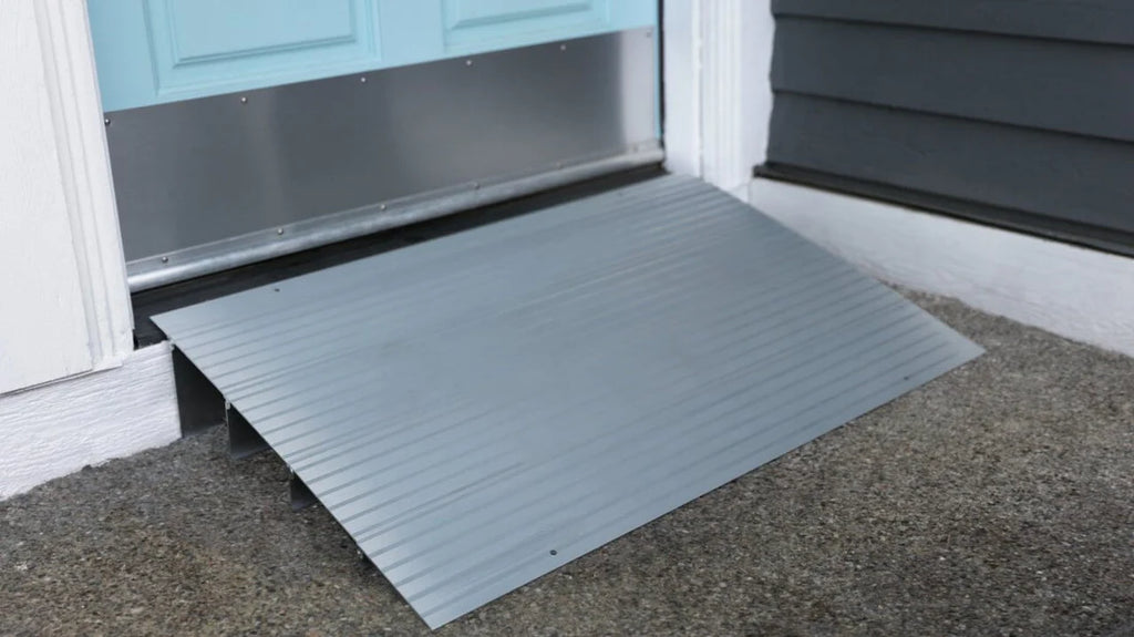 An EZ-Access TRANSITIONS® Modular Entry Ramp with a slip-resistant surface is installed at the house entrance, providing wheelchair access over the doorstep. The door is light blue, and the exterior wall is dark gray with white trim.