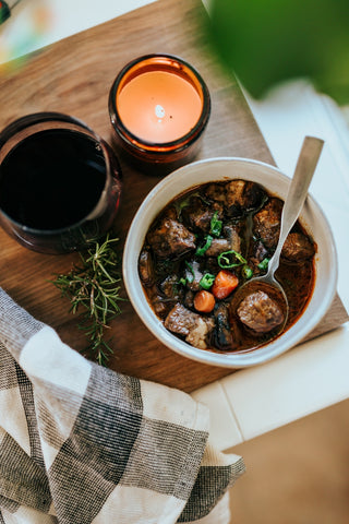 A bowl of dark broth-based soup with carrots, green garnish, and meatballs. Candles and a glass of red wine sit next to the bowl.