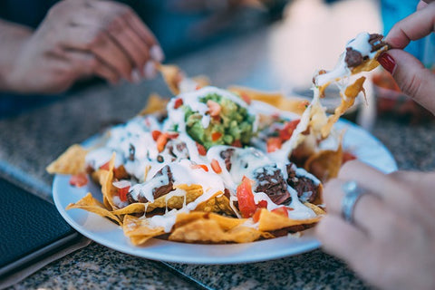 Three hands reaching in for a plate of nachos on a white styrofoam plate.