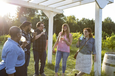 A group of 5 visitors enjoying a glass of wine under the sunlight and in lush green surroundings.