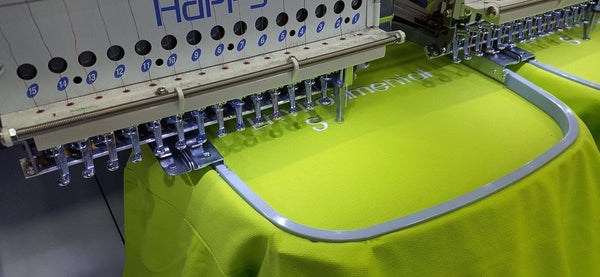 Close up of the embroidery machines, a lime green polo is being embroidered with white thread spelling out enviromental services