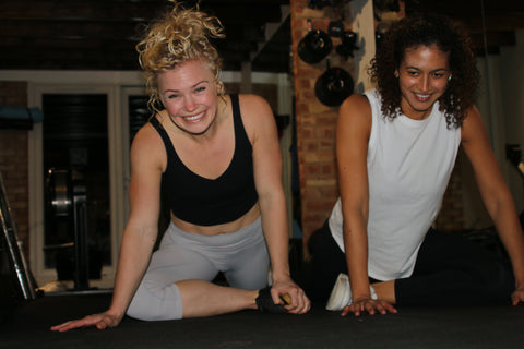 Two CrossFit girls performing a hurdle stretch in a home gym.