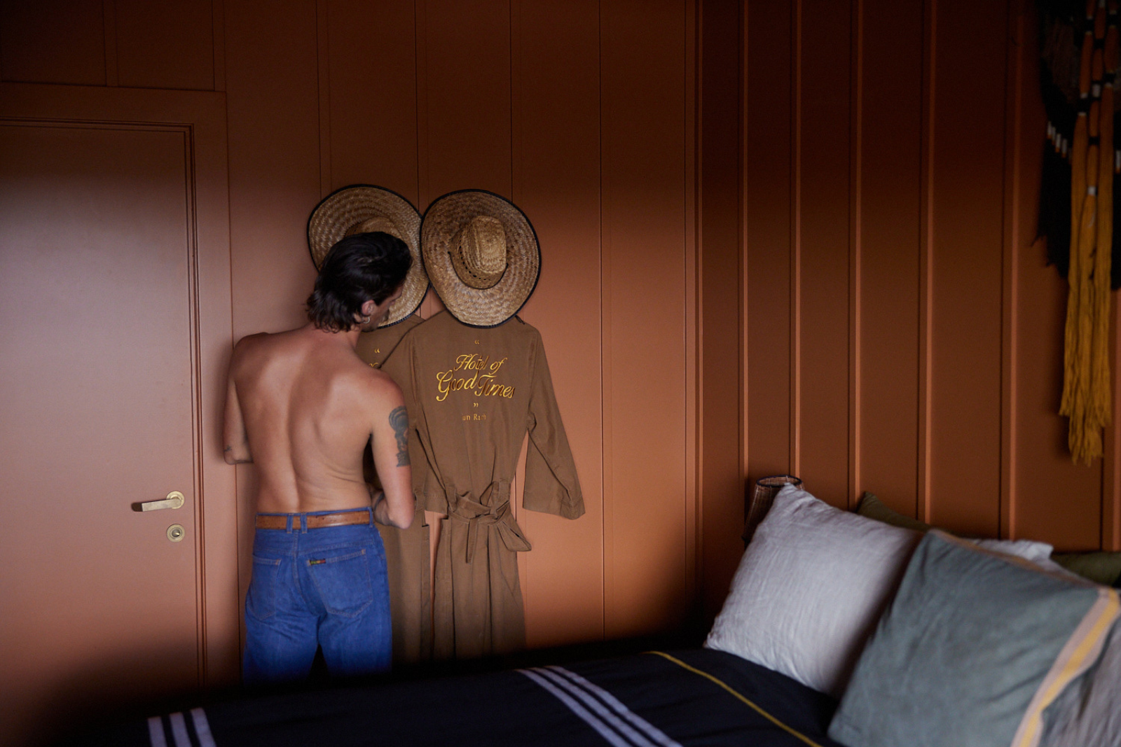 Man with jeans and no shirt facing a burnt orange wall with two tan cotton robes and two cowboy hats hanging on it.