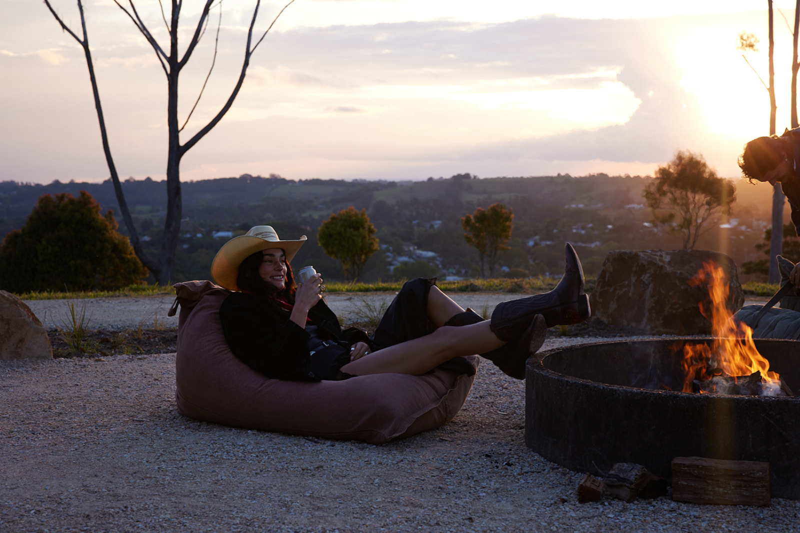 Woman in 20's laid back on bean bag by outdoor fire pit with sunset in background