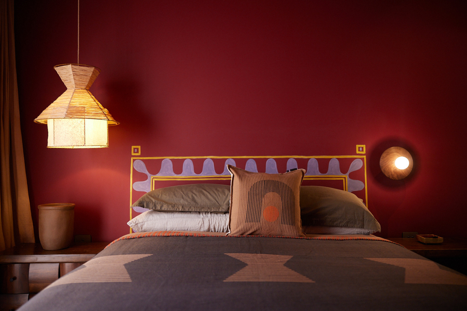 Queen Bed with aztec patterned cushion and throw. Room has dark red wall and golden lampshade on left. Room is very moody
