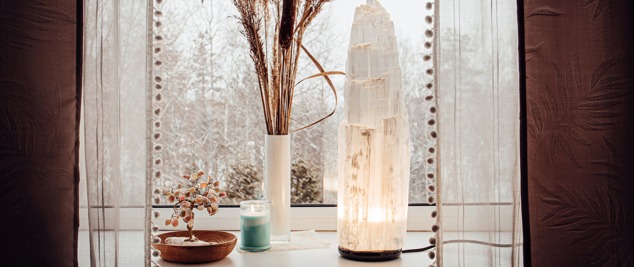 On a windowsill, curtains open, there is a Selenite Lamp, Citrine tree, and potted pampas grass