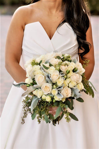 A bride in a white dress holds a bouquet of white roses and greenery, focusing on the bouquet with the bride's face not shown.