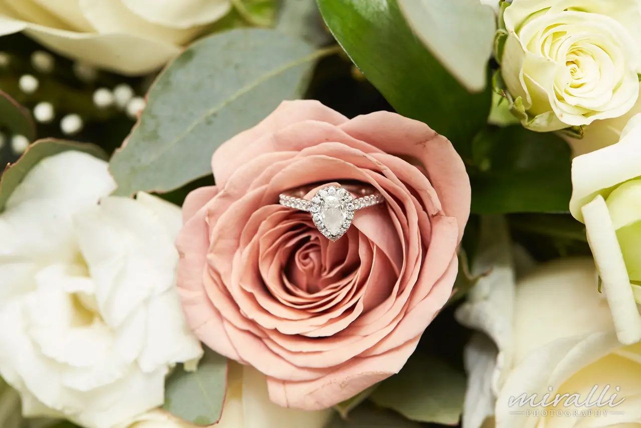 Close-up of a pink rose with a sparkling diamond engagement ring nestled in its petals, surrounded by white roses and green leaves, with a blurred background.