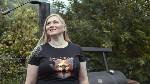 Maja steh vor einem Amerikanischen BBQ-Smoker im Garten. Sie trägt ihr Wildspark Signature Shirt aus nachhaltiger Bio-Baumwolle. Das Design verkörpert die Stärke der Frauen, ihre Leidenschaft die Feuerwehr und die Liebe zum Grillen sowie das Feuer selbst.
