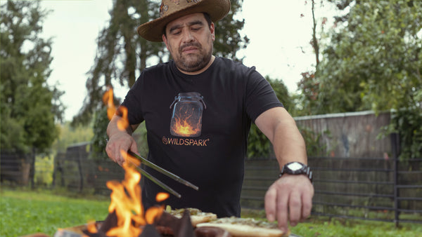 Hawk grillt an der Feuerplatte im Garten. Er trägt ein nachhaltiges Shirt aus Bio-Baumwolle von Wildspark mit dem Design Spark Jar.