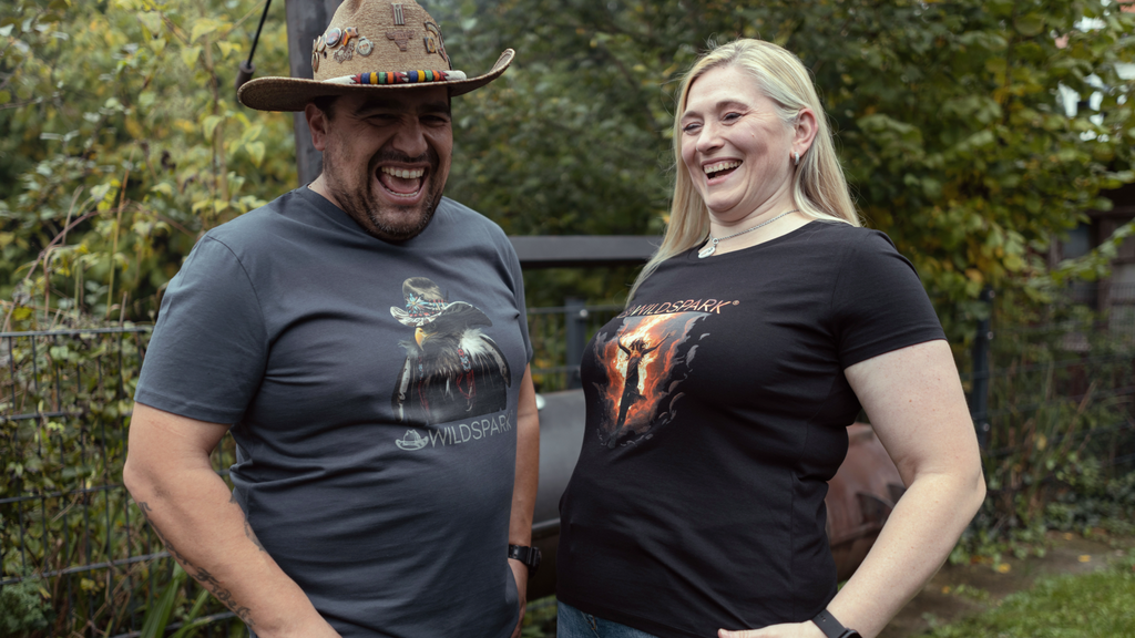 Die Brand Ambassadors von Wildspark, Maja Weigt und Hawk Blackburn in ihren Signature Shirts. Sie stehen lachend vor einem BBQ Smoker.