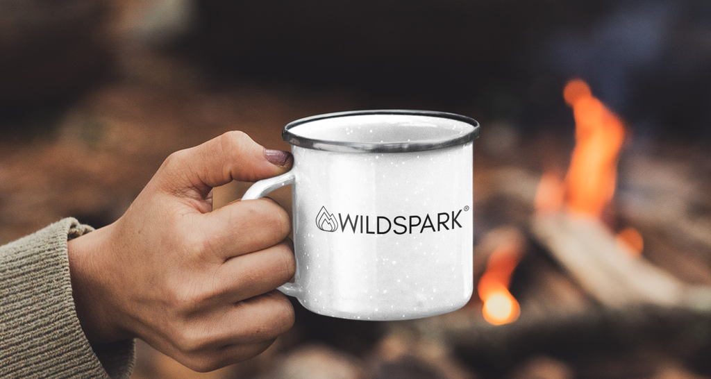 Wildspark Emaille Tasse in weiß mit Logo und Brandname. Wird von einer Hand vor einem Lagerfeuer gehalten