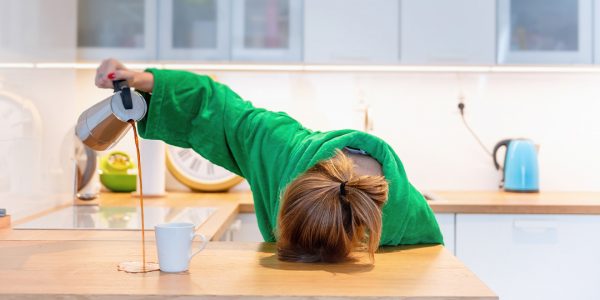image of a woman lying her head on the kitchen counter in tiredness, pouring coffee into a white mug but missing the mug