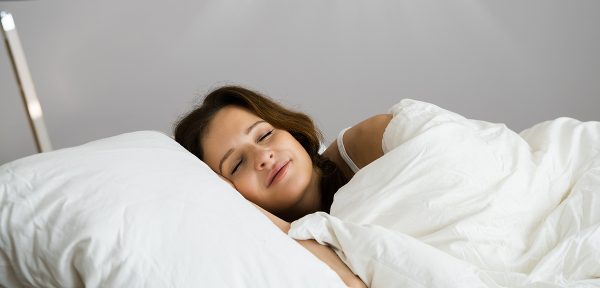 a woman with brown hair lies in bed on a white pillow and a white blanket, she looks happy and cosy