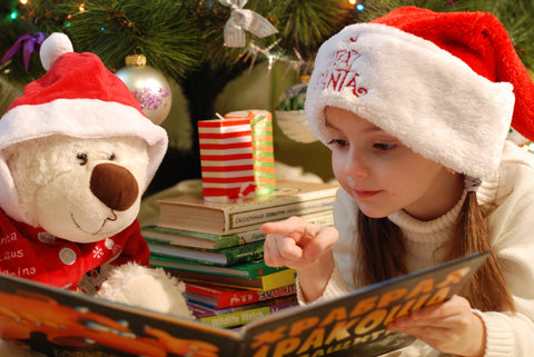 little girl wearing a santa hat reading a book to teddy bear in front of christmas tree